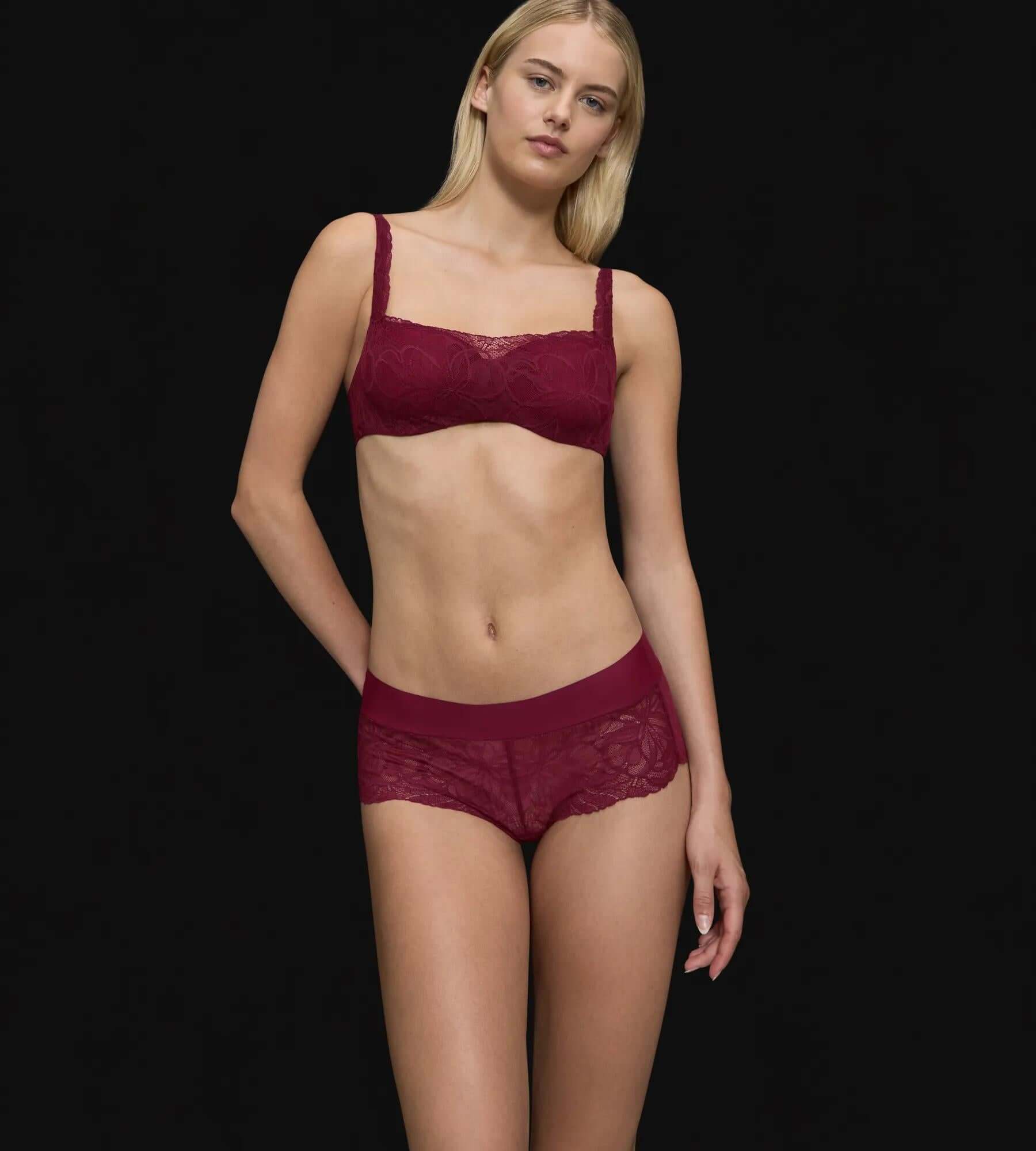 Model wearing Body Make-Up Illusion Lace shorty briefs and matching bra in burgundy against a black background.